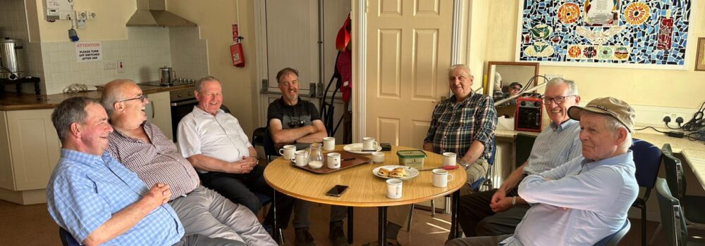 Group of elderly men sitting around a table in the Men's Shed drinking tea and eating cake