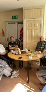 Group of elderly men sitting around a table in the Men's Shed drinking tea and eating cake