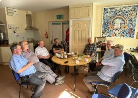 Group of elderly men sitting around a table in the Men's Shed drinking tea and eating cake