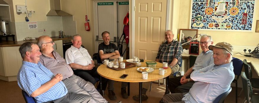 Group of elderly men sitting around a table in the Men's Shed drinking tea and eating cake