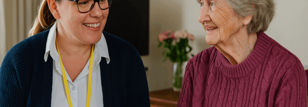 A female occupational therapist in a bright home setting smiles as she speaks with an elderly woman seated comfortably in a chair, reflecting supportive care and community-based therapy.