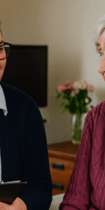 A female occupational therapist in a bright home setting smiles as she speaks with an elderly woman seated comfortably in a chair, reflecting supportive care and community-based therapy.