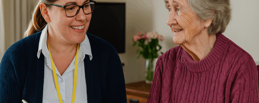 A female occupational therapist in a bright home setting smiles as she speaks with an elderly woman seated comfortably in a chair, reflecting supportive care and community-based therapy.